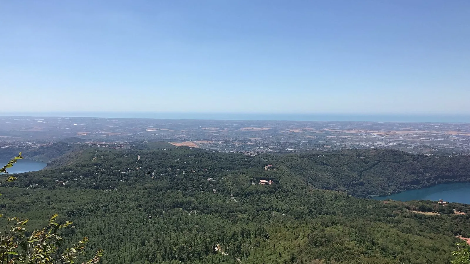 Veduta dall'alto dei Colli Albani boscosi con due laghi vulcanici ai lati e, sullo sfondo, la pianura costiera e il mare